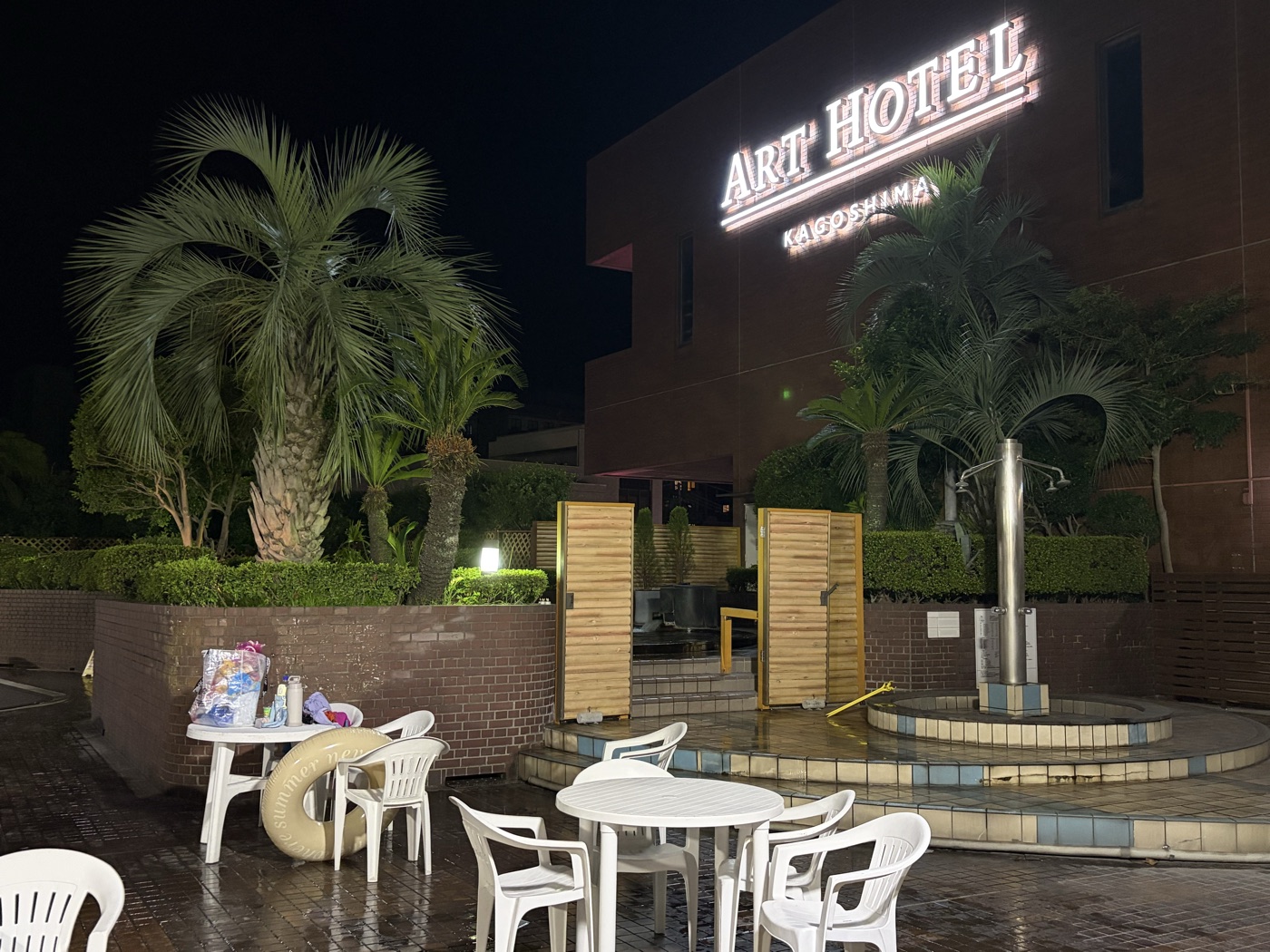 夜のアートホテル鹿児島のガーデンプールサイド。ライトアップされたヤシの木と白いテーブル、奥にART HOTEL KAGOSHIMAのネオンサイン